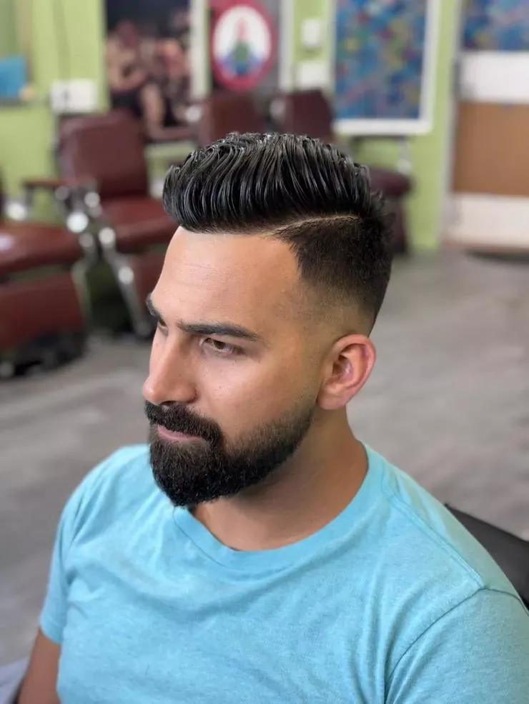 Side view of a man with a high fade, combed top and full beard in a barber chair